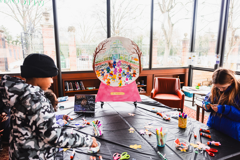 kinds doing crafts at a table in a room with large glass windows showing a yard and trees behind them