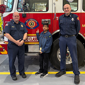 two male firefighters in blue uniform stand by fire truck with a boy pointing to the truck