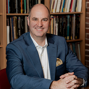 man smiling in blue suit with shelves of books in background
