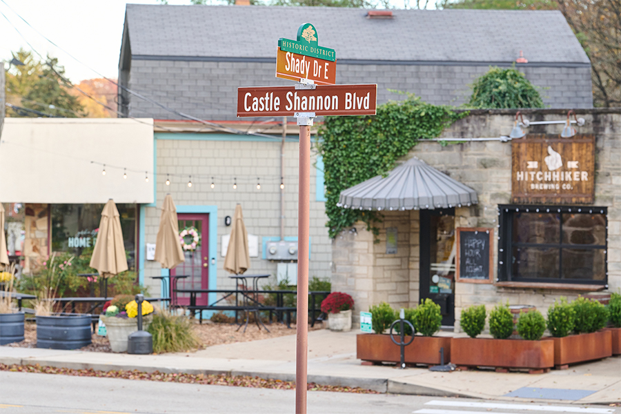 A street sign in the historic district with the words Shady Dr E and Castle Shannon Blvd in white letter. A building with ivy and patio lights and umbrellas can be seen in the background.