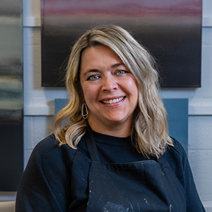 A woman with shoulder length blonde hair wearing a black long sleeve top and a black apron with a cheerful expression sitting in a chair in front of a gallery wall filled with abstract paintings.