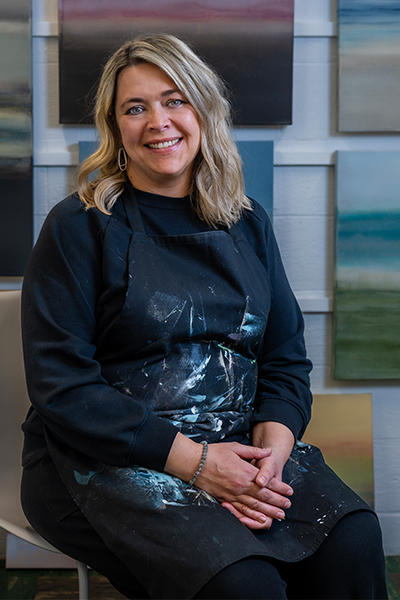 A woman with shoulder length blonde hair wearing a black long sleeve top and a black apron with a cheerful expression sitting in a chair in front of a gallery wall filled with abstract paintings.