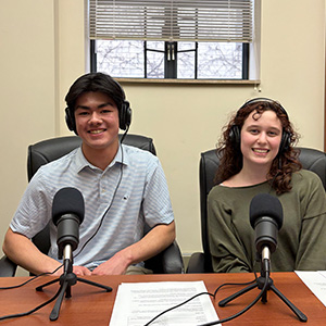 a teen boy and girl sit behind microphones wearing headphones