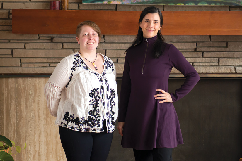 two adults standing in front of a stone wall, one wearing a black and white shirt and pants and the other wearing a purple dress