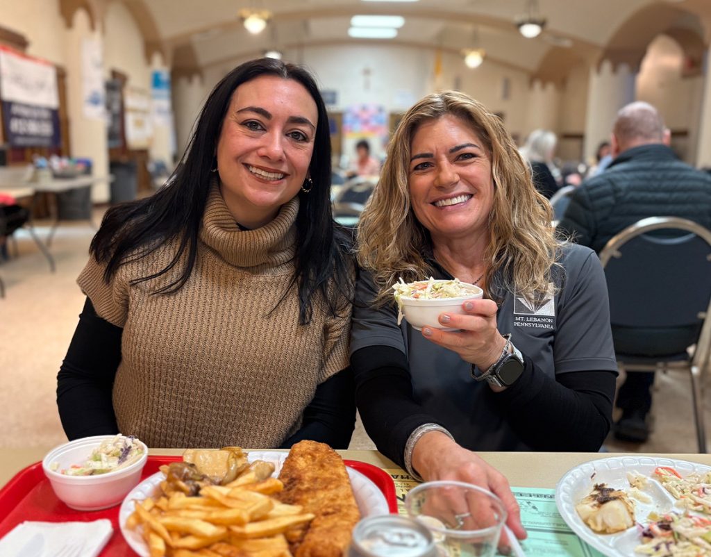 Two women smiling as they are seated at a table with food in front of them. The woman on the left is holding up coleslaw.