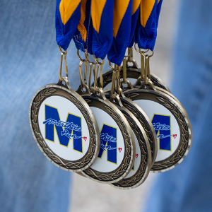 A close up shot of gold medals with a large white circle and the letter M in the middle attached to blue and yellow ribbon.
