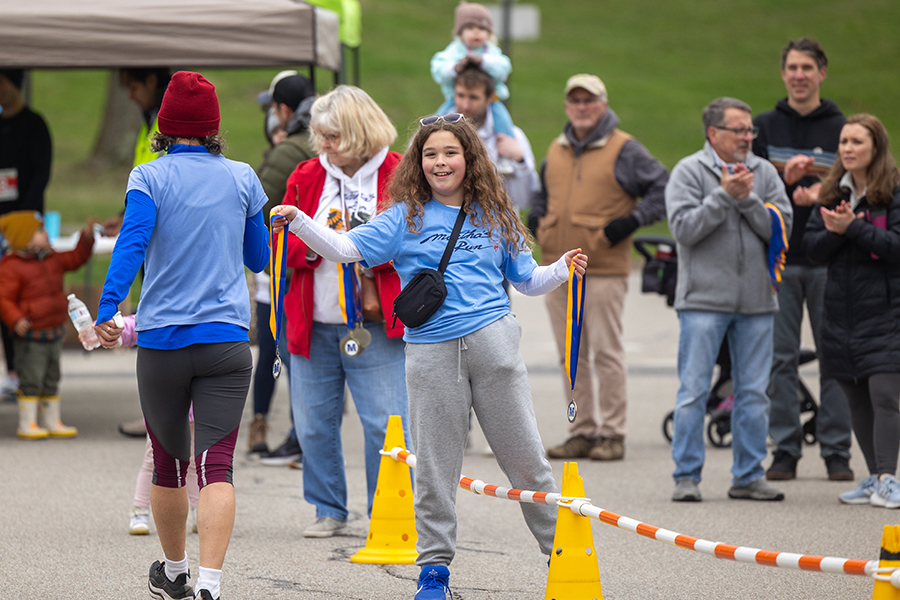 A tween in a blue shirt and gray pants standing over yellow cones holding medals smiles for the camera. A runner and spectators can be seen in the background.
