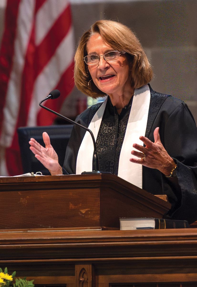 a female preacher standing at a a podium with a microphone speaking. her arms are spread and she is wearing a black robe with a white scarf