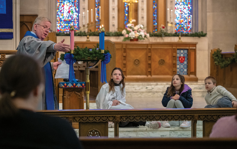 A male preacher speaks to a congregation in the pit, wearing robes, while three young girls sit on the steps watching