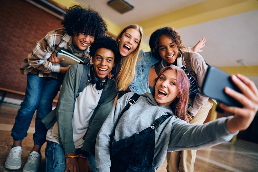 Group of cheerful high school students taking selfie with cell phone in hallway.