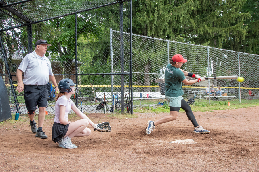 An action shot of a softball game with a player in a green shirt and red hat hitting a softball with the catcher and umpire behind home plate. Green trees, a chain link fence and spectator benches can be seen in the background.