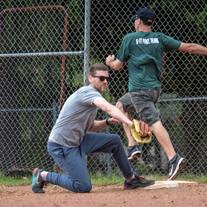 An action shot of a softball game with a player in a gray shirt and blue pants catching a softball with one foot on the base. Another player can be seen running in the background.