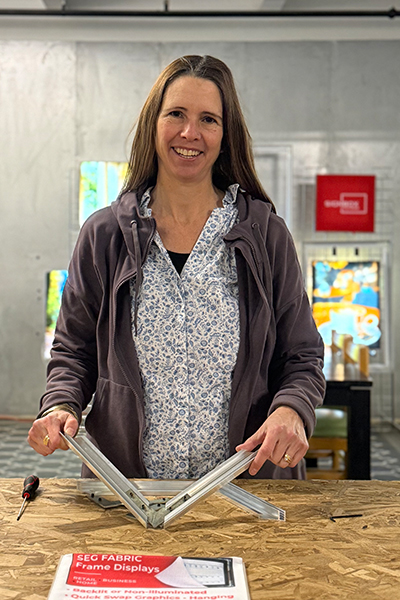 A woman with long brown hair wearing a blue and white button down top and a purple sweatshirt smiles while holding small metal frame pieces.