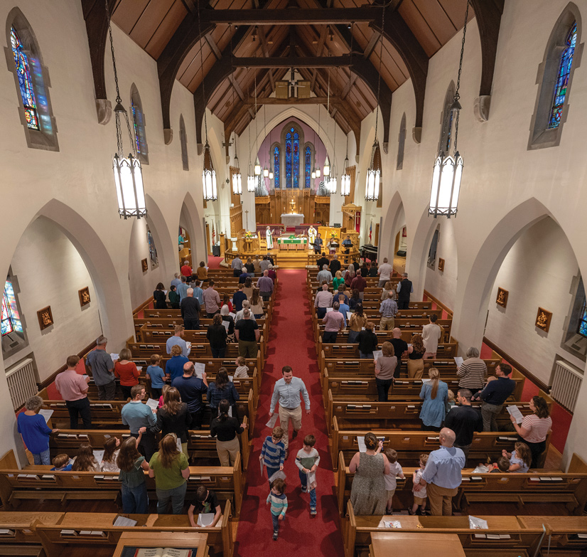 the inside of st. pauls episcopal church, there is a tall cieling, large windows, people are standing in the pews while the preacher stands at the alter speaking