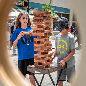 Two boys one with wearing a bright blue tshirt and black shorts and the other wearing a gray shirt and a baseball hat playing an over-sized tower Jenga tower game.