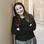 young woman in black long sleeve shirt with arms crossed leaning on a tan wall smiling with long brown hair