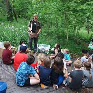group of children sitting on a blanket in a wooded park with a teenager in a flannel teaching them