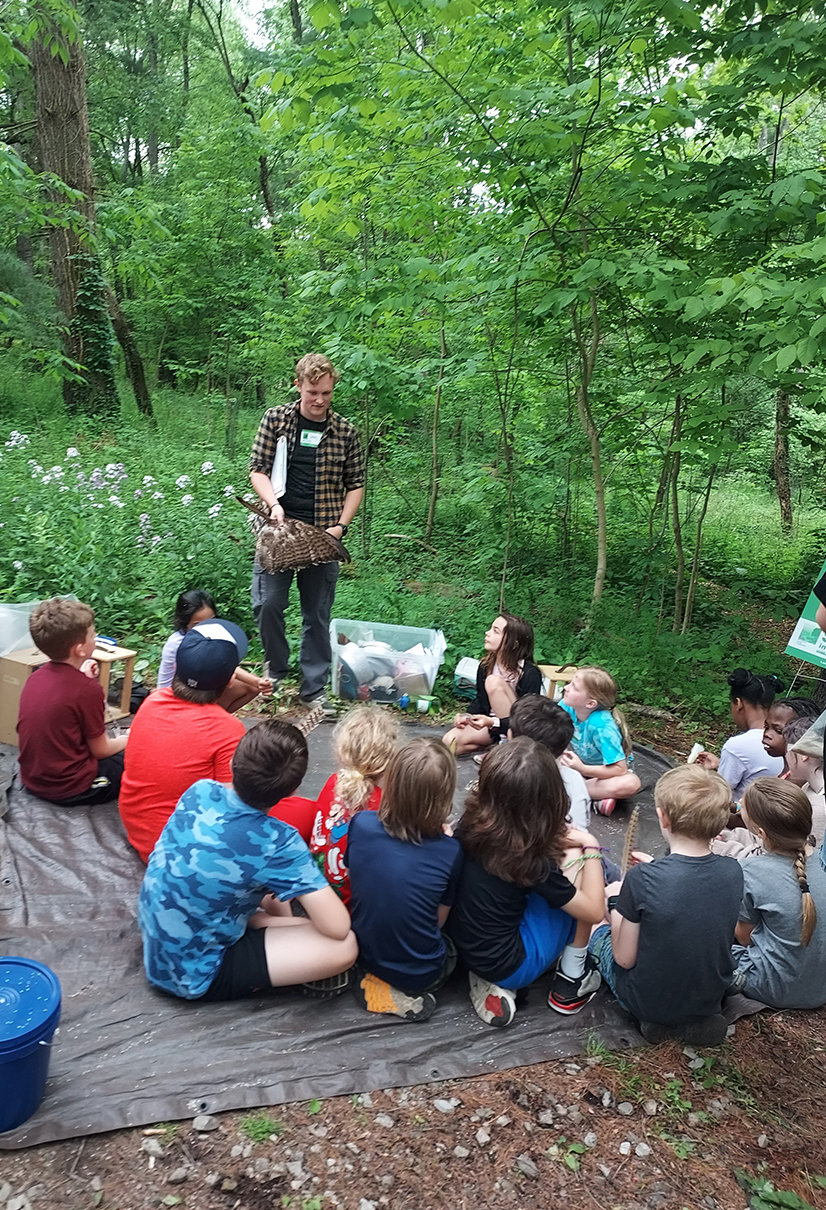 group of children sitting on a blanket in a wooded park with a teenager in a flannel teaching them