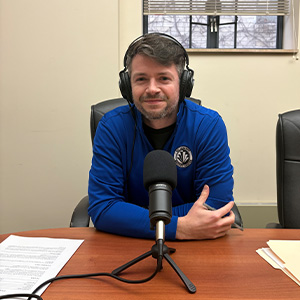 Man with blue quarter zip, wearing headphones, sits in front of a microphone