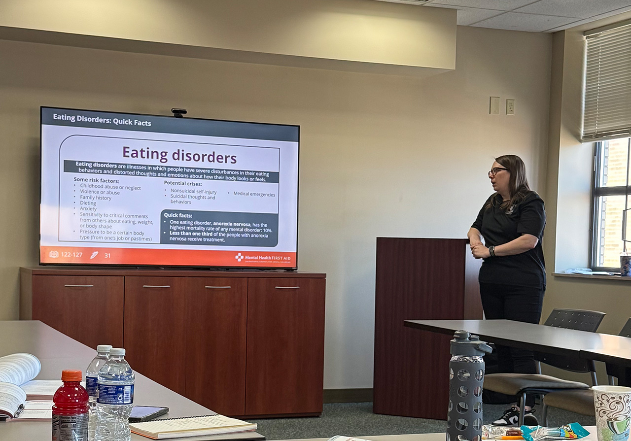 woman in black pants and black polo stands beside a podium presenting a slide on eating disorders