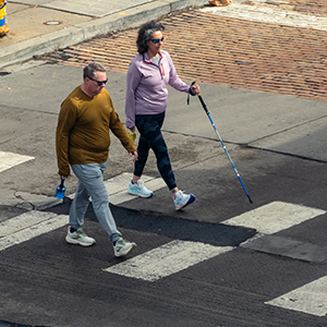 two adults walking in a crosswalk, one holding a walking stick