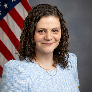 headshot of woman in blue top with american flag in background