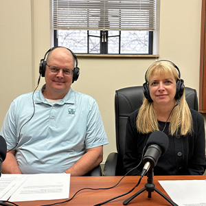 A man in a blue polo and a woman with long blonde hair wearing a black long sleeved top sitting next to each other. Both are wearing headphones and are sitting in front a microphone.