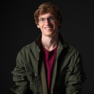 headshot of man with glasses and green jacket on black background