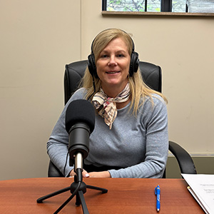 Woman with grey long sleeved top and scarf sitting in front of a microphone with headphones